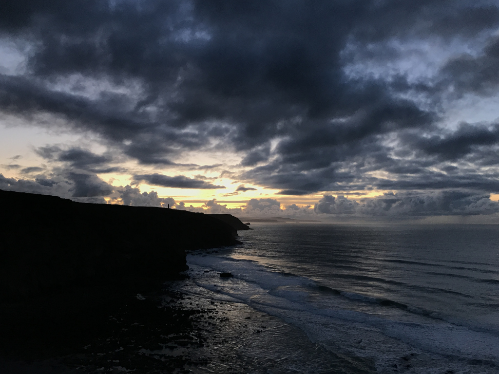 Dramatic coastal scene at sunset or sunrise. A dark, silhouetted cliff dominates the left side of the frame, while the right shows a wide expanse of ocean with gently rolling waves. The sky is a mix of dark, brooding clouds and lighter, brighter areas where the sun is breaking through, creating a contrast between light and shadow. A solitary figure can be barely made out on the cliff's edge, adding a sense of scale and isolation. The overall mood is moody, atmospheric, and somewhat melancholic.
