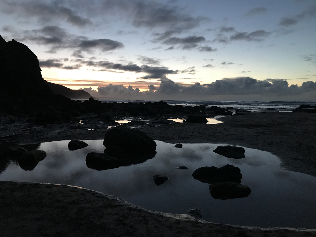 Tranquil coastal scene at twilight. Dark, silhouetted rocks dominate the foreground, surrounding a small pool of water that reflects the muted colours of the sunset sky. The ocean stretches out in the background, under a sky filled with clouds exhibiting a range of colours from deep blues to warm oranges and yellows. The overall mood is serene and somewhat melancholic, emphasising the quiet beauty of the natural landscape.