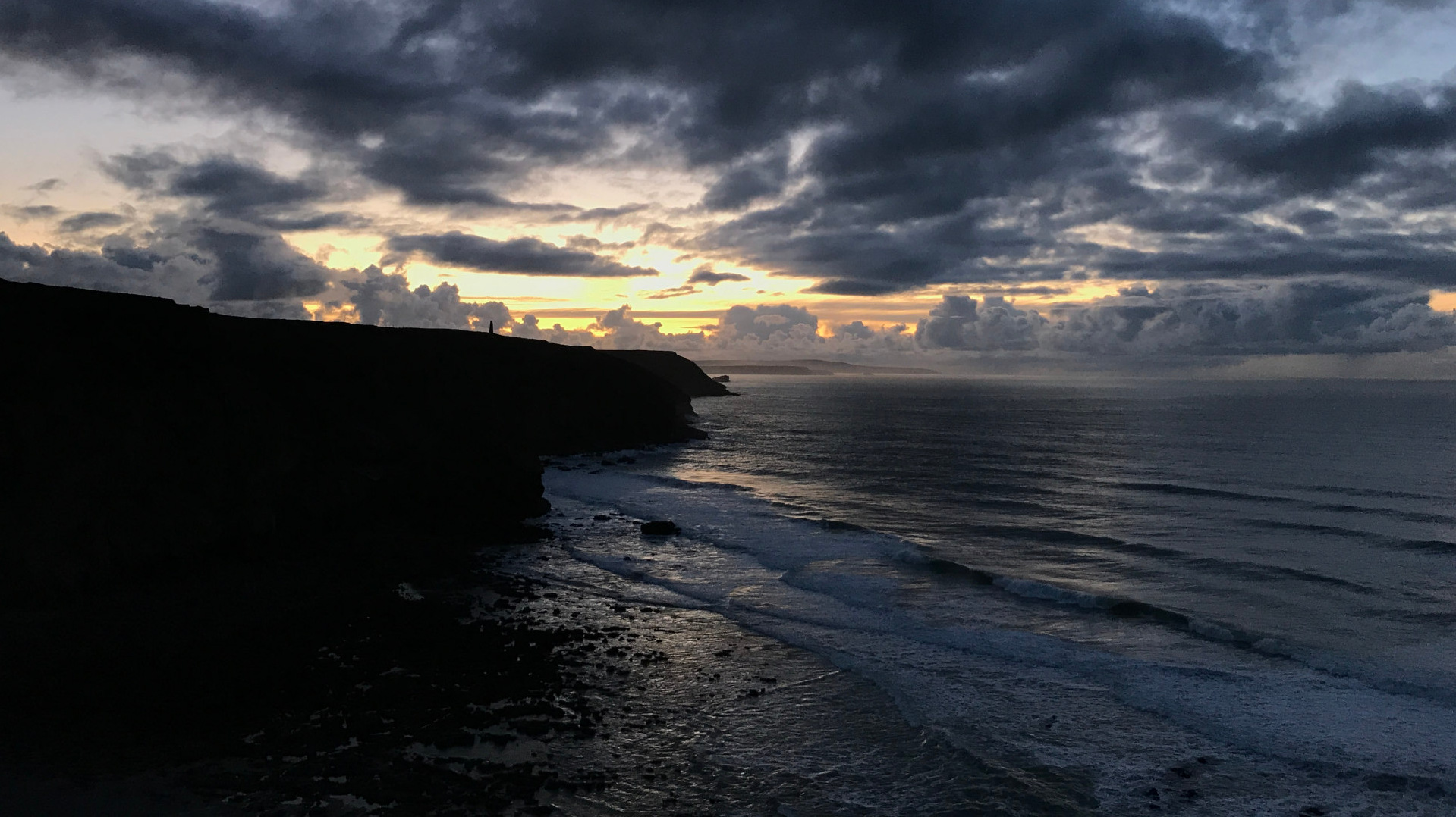 A dramatic coastal scene at either dawn or dusk. A dark, silhouetted cliff face dominates the left side of the frame, contrasting sharply with a vast, moody ocean extending to the horizon. The sky is a breathtaking display of dark, brooding clouds, punctuated by streaks of pale yellow and orange light breaking through, suggesting a sunset or sunrise. The overall mood is one of powerful, raw beauty and a sense of impending change or transition.