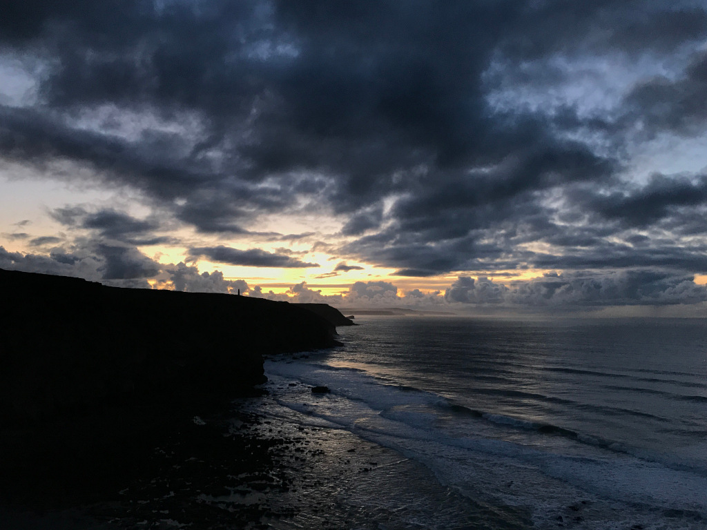 Dramatic coastal scene at either dawn or dusk. A dark, silhouetted cliff face dominates the left side of the frame, contrasting sharply with a vast, moody ocean extending to the horizon. The sky is a breathtaking display of dark, brooding clouds, punctuated by streaks of pale yellow and orange light breaking through, suggesting a sunset or sunrise. The overall mood is one of powerful, raw beauty and a sense of impending change or transition.