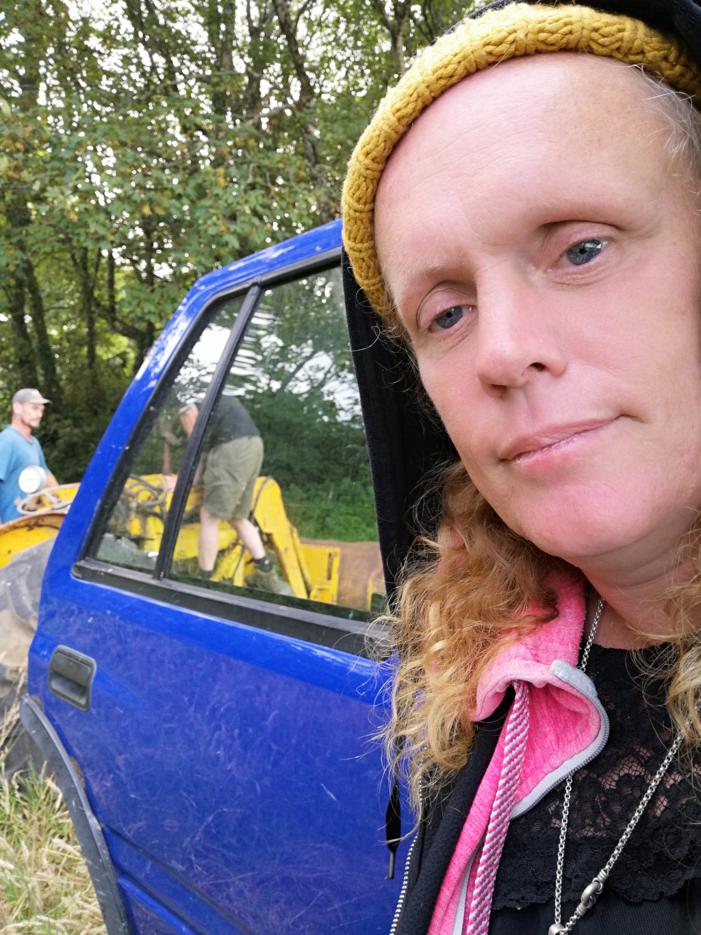 Close-up selfie of Leonie with shoulder-length, wavy blonde hair. She's wearing a dark-coloured hooded sweatshirt with a mustard-yellow knitted beanie pulled low over her forehead. A vibrant pink fleece neck warmer or scarf is partially visible around her neck, and she has two silver necklaces. Her expression is serious, almost pensive, with a slightly downturned mouth. Her skin is fair with visible freckles or blemishes. Behind her, out of focus but still visible, is a bright blue, older-model SUV. Partially visible through the SUV's back window are two men working on a yellow tractor or excavator. One man, wearing a baseball cap, seems to be observing; the other, wearing a lighter-coloured shirt and shorts, appears to be operating the machinery.