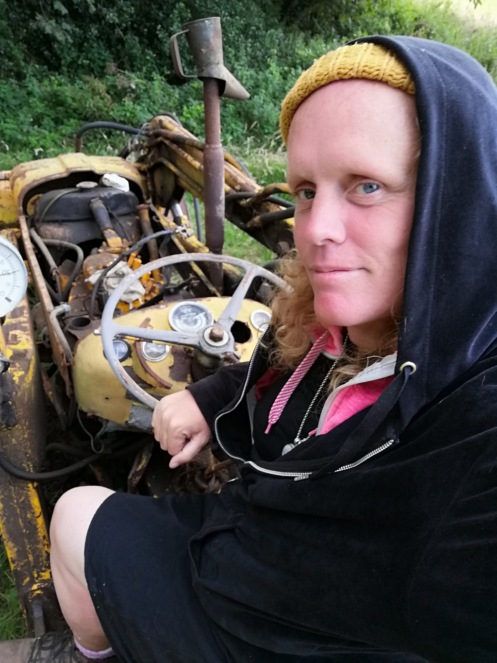 Focal point is a fair-skinned woman with shoulder-length, wavy auburn hair, partially concealed by a dark navy blue hoodie with a drawstring hood. She's wearing a mustard yellow knit beanie hat and a dark-coloured, possibly black, short skirt or shorts. A pink drawstring hoodie is visible underneath the navy one. She's seated behind the wheel of a rusty, yellow vintage tractor. Her right hand rests on the steering wheel while her left hand gently grips the gear shift. She gazes directly at the camera with a neutral to slightly pensive expression. Her facial features are soft, her lips are slightly parted, and her brow is unfurrowed. A silver necklace is visible around her neck. The tractor itself is old and weathered. Its yellow paint is chipped and faded, revealing rust in various places. Its mechanical components are partially exposed, showcasing wear and age. The steering wheel is worn, and various gauges and controls are visible on the dashboard. A rusty yellow bucket arm extends out from the tractor's chassis.