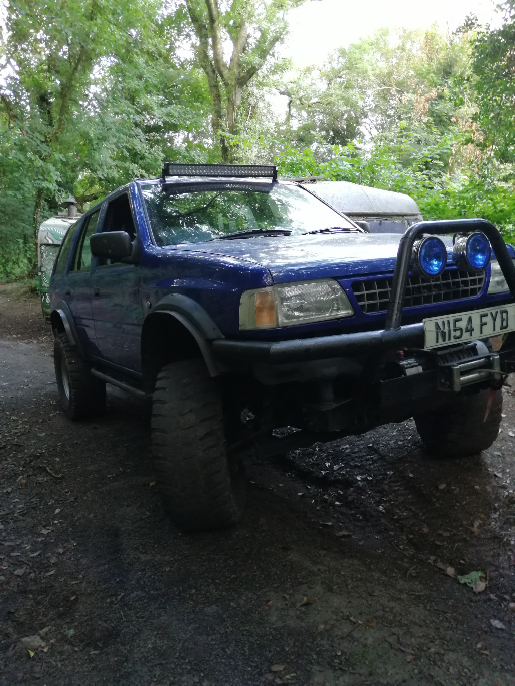 Modified, blue Opel Frontera Sport SUV. It's significantly lifted, boasting oversized, heavily tread all-terrain tires that are dark gray-black and heavily soiled. A substantial, black bull bar with integrated round blue auxiliary lights dominates the front. Atop the wind shield sits a long, straight LED light bar, adding to its off-road aesthetic. The vehicle is parked on a slightly uneven, dark gray-brown gravel or dirt road, angled slightly away from the viewer, allowing for a partial side and front view. The license plate reads N154 FYB. Partially visible in the background, slightly out of focus, is a light-coloured caravan or camper trailer, suggestive of camping or outdoor adventure. The setting is a wooded area, with lush green foliage forming a natural backdrop. Tall trees, some showing thick trunks and branches, surround the vehicle. The lighting suggests it's likely daytime, possibly late afternoon or early evening, as the light is relatively soft and diffused, casting few harsh shadows.