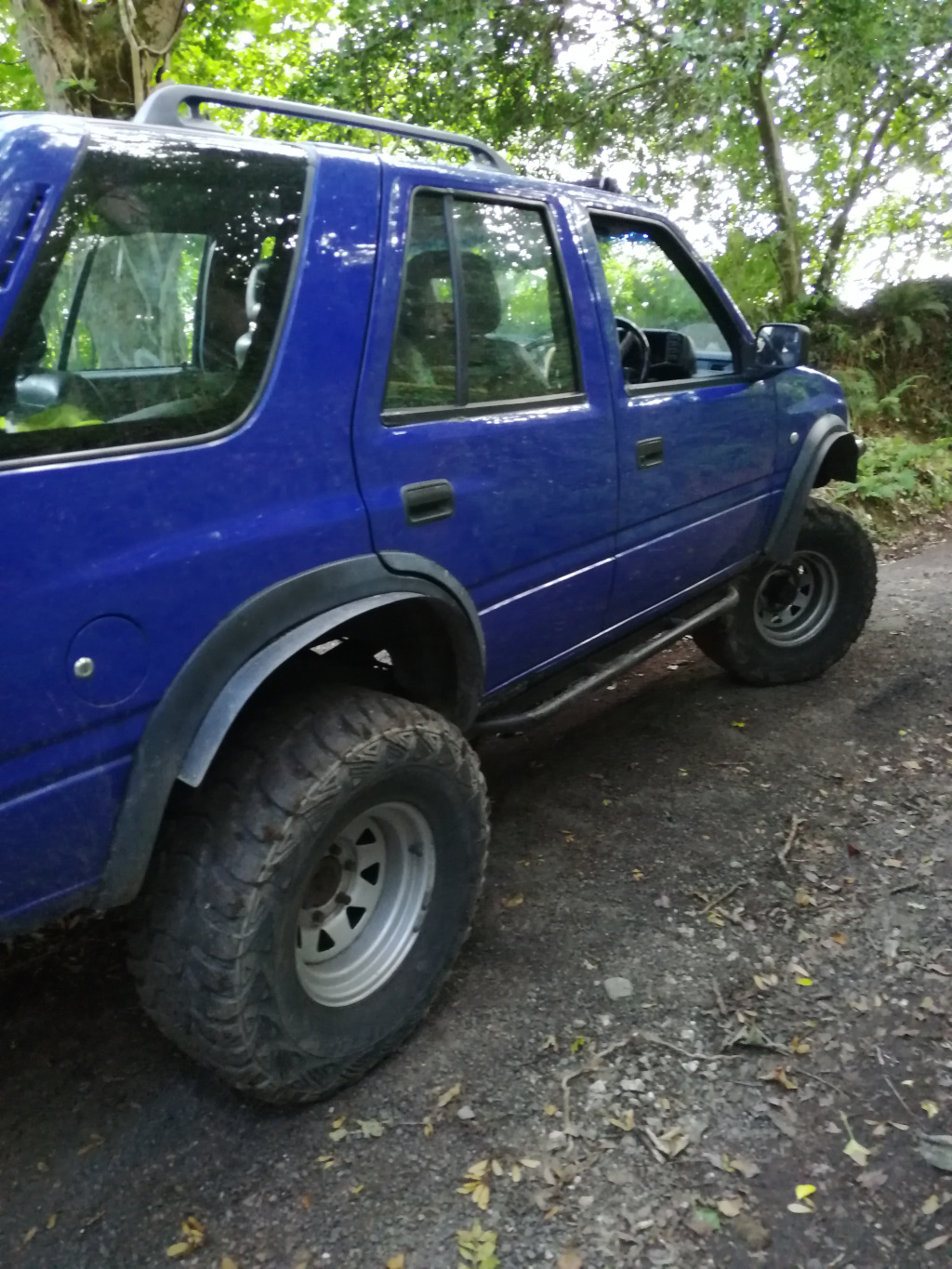 Vibrant, royal blue Opel Frontera Sport, a four-door SUV, modified for off-roading. It's parked on a gravel road, angled slightly away from the viewer, showcasing its right side. The vehicle is lifted, boasting large, aggressive all-terrain tires that fill the wheel wells.  The tires have a deep tread pattern, showing signs of wear consistent with off-road use. The vehicle is equipped with aftermarket side steps or running boards, adding to its rugged appearance. A person is visible behind the driver's-side window, though details are obscured by the glass and the angle. The roof rack is clearly visible.