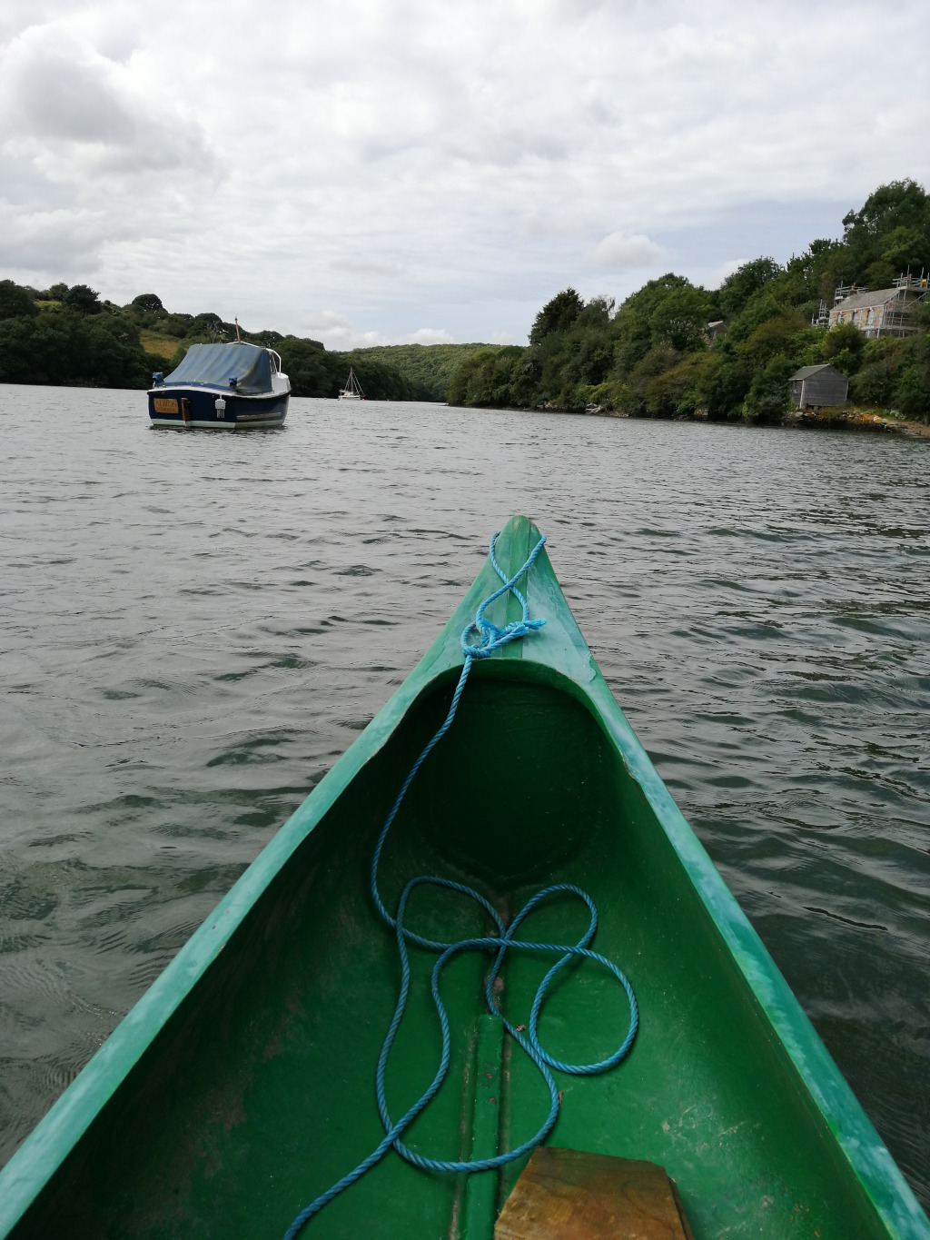 First-person perspective from inside a green canoe paddling down a calm river. Two other boats are visible in the distance, one larger covered boat and a smaller sailboat, nestled among gently sloping green banks. The sky is overcast. The overall mood is peaceful and serene.