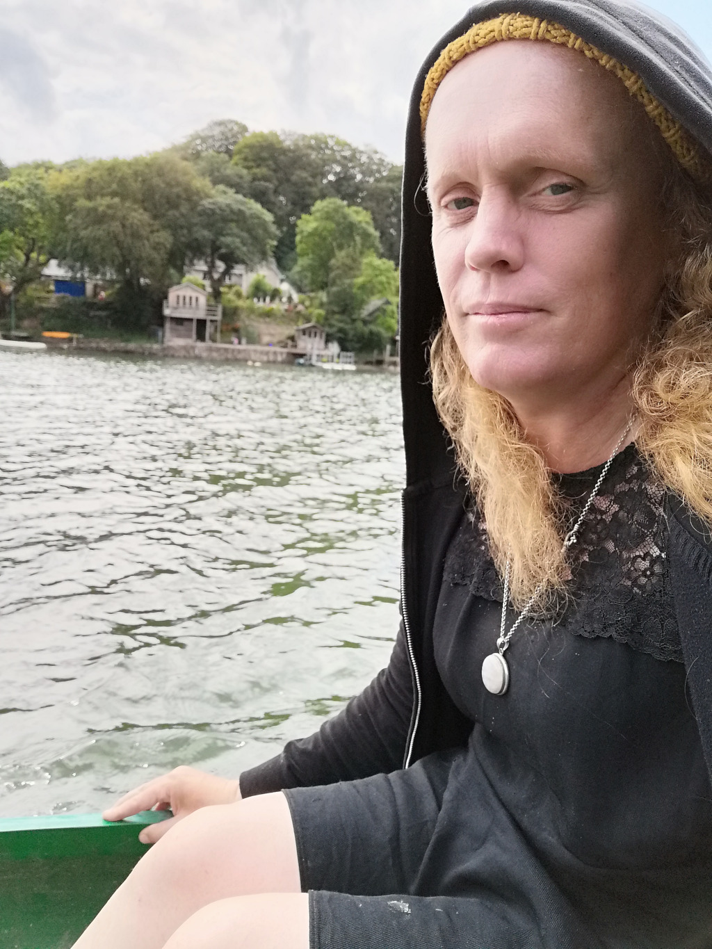 Middle-aged woman with shoulder-length blonde hair, wearing a black hooded jacket and a black dress with lace detailing. She's seated in a small green boat on a lake. In the background, there's a slightly blurry view of houses nestled among trees on the lakeshore. The overall mood is calm and contemplative.