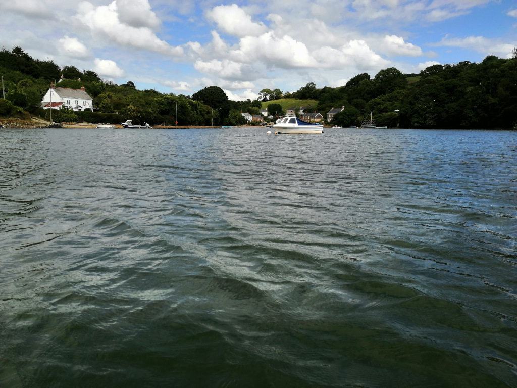 Calm river scene. In the foreground is the dark, slightly rippled water. In the mid-ground and background is a picturesque shoreline with lush green vegetation, several houses, and a few small boats moored — one of which is prominently centered in the mid-ground. The sky is partly cloudy, with a mix of white fluffy clouds and blue sky. The overall impression is one of tranquillity and peaceful natural beauty.