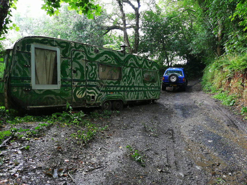 Green, elaborately painted caravan parked on a muddy, overgrown track in a wooded area. A blue SUV is parked further down the track, suggesting a journey or arrival at a secluded location. The scene evokes a sense of adventure, perhaps hinting at a nomadic lifestyle or off-grid living. The caravan's unique artwork adds a layer of artistic expression to the overall setting.