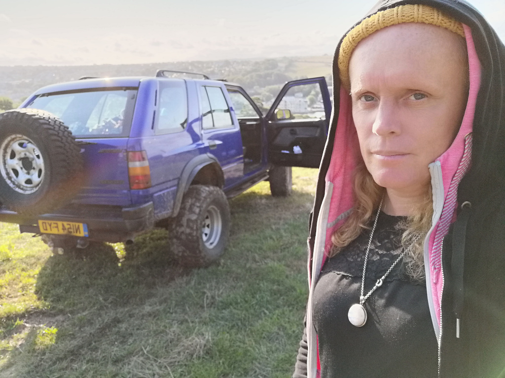 Leonie wearing a black hoodie with a pink interior, and a yellow beanie underneath. She stands in a grassy field with a blue four-wheel-drive vehicle behind her. The vehicle is parked, and its back is facing the camera. The woman's expression is serious and she is wearing a necklace. The overall scene suggests a moment of quiet contemplation or solitude in a rural setting.