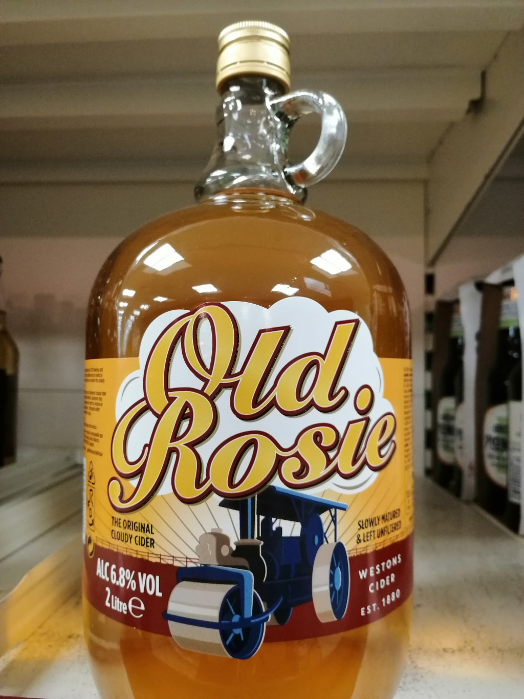 Large glass bottle of Old Rosie cider. The bottle is amber-coloured, filled with the cider, and features a vintage-style label with the brand name in a stylized script font. The label also includes an illustration of a steamroller, suggesting a traditional or nostalgic aesthetic. The text on the label indicates that it's the original cloudy cider, slowly matured and unfiltered, from Westons Cider, established in 1880.