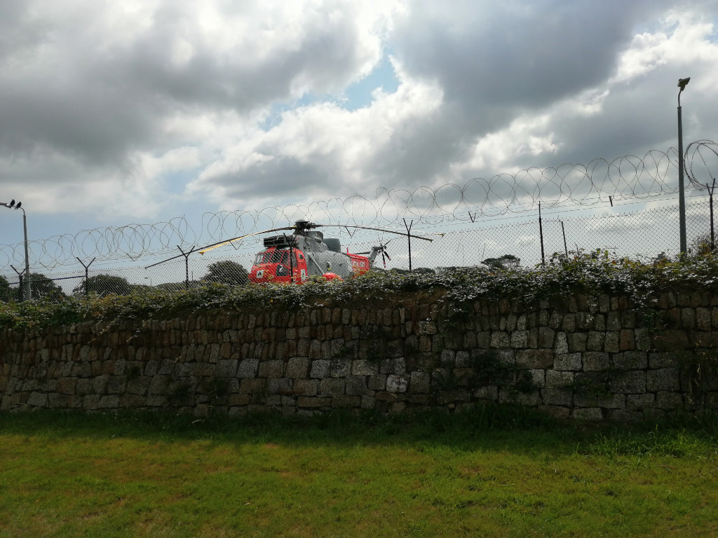 Red and grey helicopter partially visible behind a high stone wall topped with barbed wire fencing. The sky is cloudy. The scene appears to be at a secure or restricted location.