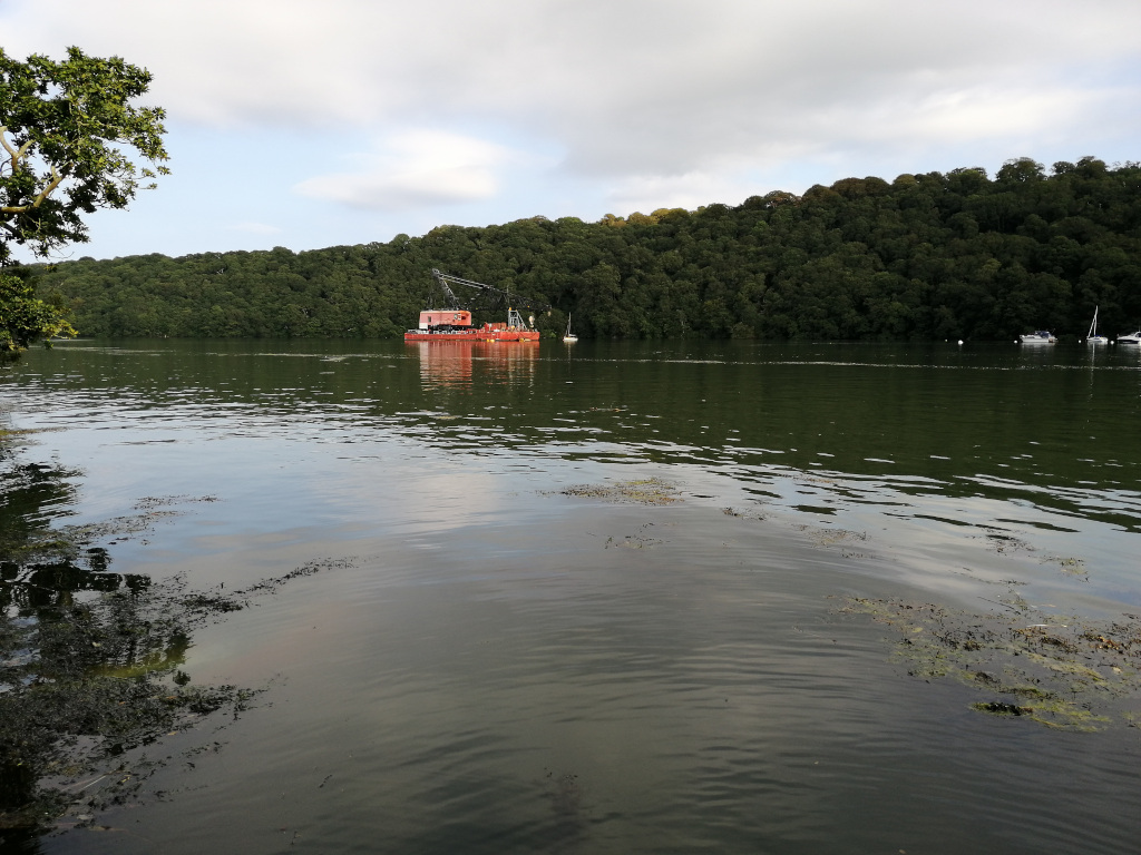 Calm body of water, possibly a river or lake, with a small red barge or work boat in the middle ground. The background features a lush, green hillside densely covered with trees. Several small boats are visible in the distance along the opposite shore. The overall scene is peaceful and tranquil, with a slightly overcast sky.