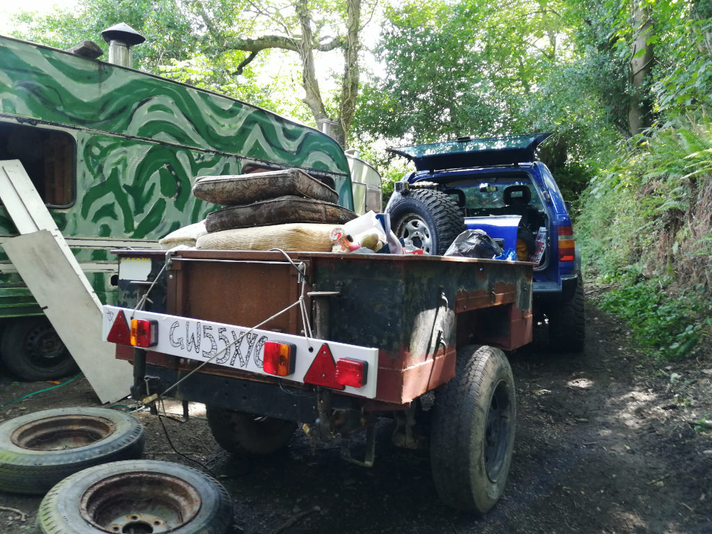 Blue SUV parked on a dirt road next to a heavily camouflaged caravan. The SUV's tailgate is open, and it's connected to a rusty, worn-out trailer loaded with various items, including what appears to be bedding and other belongings. The scene suggests a temporary or transient lifestyle, perhaps related to camping or travelling. The overall impression is one of rusticity and possibly a nomadic existence.