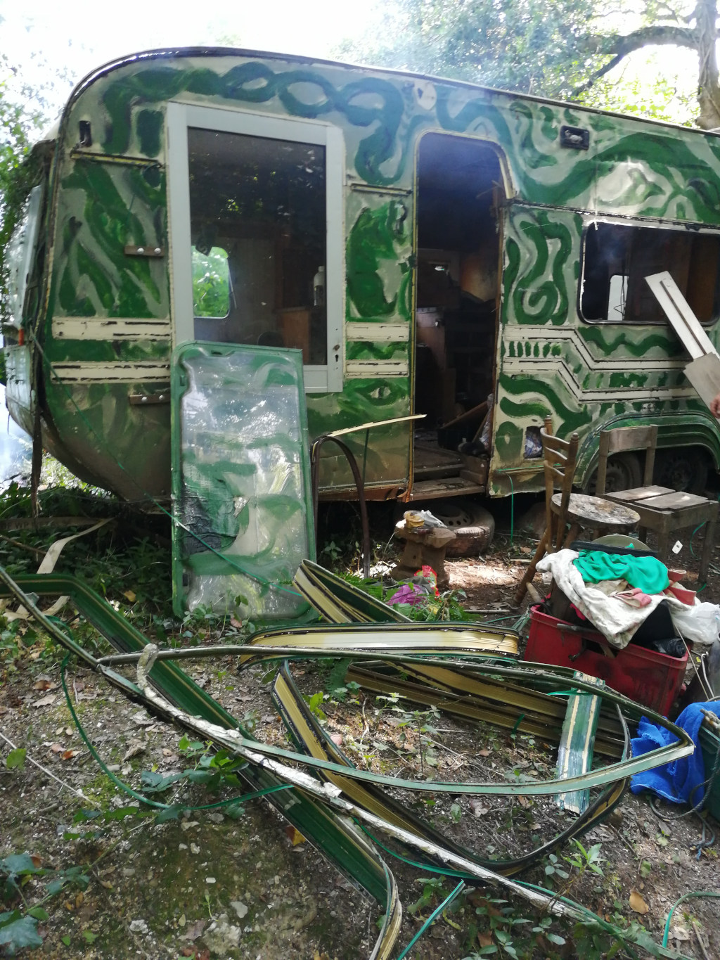 Dilapidated, green caravan, heavily adorned with abstract swirls of paint. The caravan's door is open, revealing a cluttered interior. Scattered around the caravan are broken and discarded pieces of what appears to be its window frames and other structural elements. The overall setting is a wooded area, suggesting abandonment or a state of disrepair. The scene evokes a sense of transience, perhaps hinting at a nomadic lifestyle or the remnants of a project left unfinished.