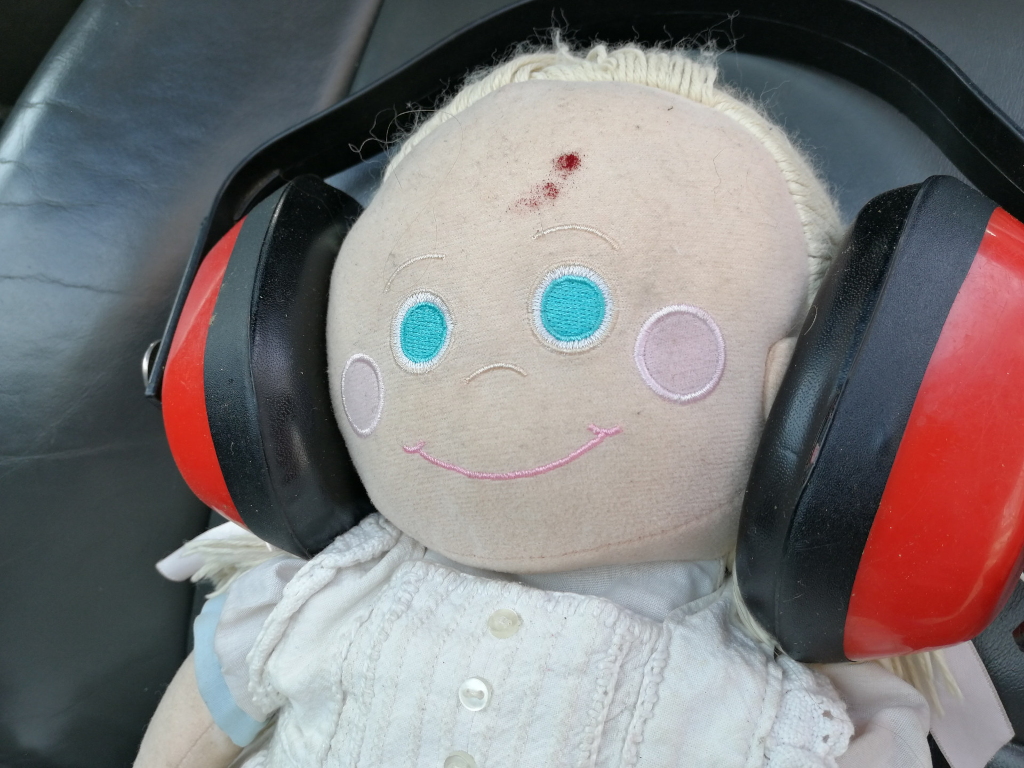 Rosie rag doll wearing a pair of red and black earmuffs. The doll has blonde hair, embroidered blue eyes, and a simple embroidered smile. It is sitting in a dark-coloured car seat, visible only partially in the background. There is a small, what appears to be a reddish-brown stain, on the dolls forehead.