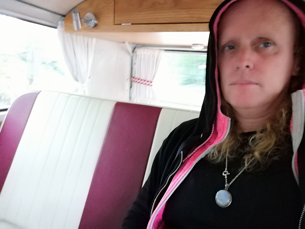Fair-skinned woman with shoulder-length blonde hair, wearing a black hoodie with pink lining, and a silver necklace with a round pendant. She is sitting in the back of a vintage camper van with white and maroon upholstery. The woman's expression is serious and neutral. The overall mood is somewhat sombre and introspective.