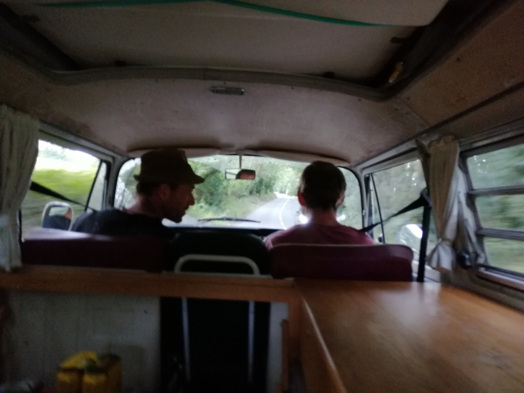 Interior of a vintage Volkswagen van, seemingly converted into a camper van. Two individuals are seated in the front, looking ahead. The van is in motion, as indicated by the blurred background of a rural road. The atmosphere is somewhat dark and moody, suggesting a journey or journey's end. The focus is on the two people and the simple, lived-in interior of the van.