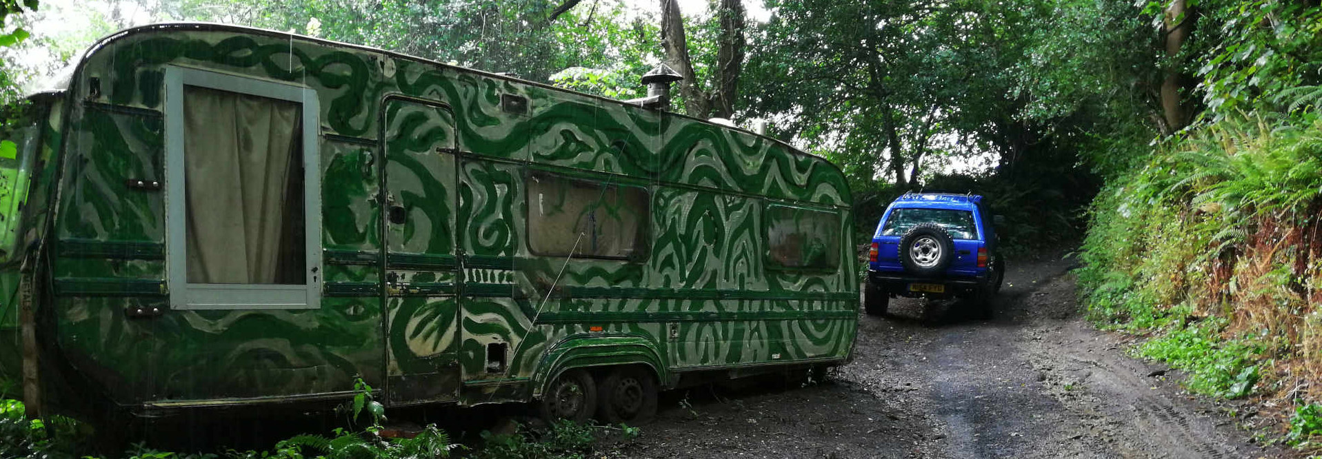 Two main objects: a distinctively painted caravan and a blue SUV. The caravan is positioned slightly off-centre to the left, angled slightly away from the viewer. Its exterior is predominantly dark green, heavily adorned with swirling, abstract, light-green and white patterns that resemble stylised vines or organic forms. The paint appears somewhat worn and textured, giving it a slightly weathered and vintage look. A small, grey window with a light-beige curtain is visible on the caravan's side. The caravan sits slightly askew, resting on its wheels. The blue SUV, appearing to be a late 20th-century model, is smaller than the caravan and positioned behind it, ascending a slightly upward-sloping dirt road. It's a vibrant, almost electric, blue, and its wheels are facing directly up the path. The tires are relatively large, indicating potential all-terrain capabilities. Both vehicles appear stationary. There are no people visible.