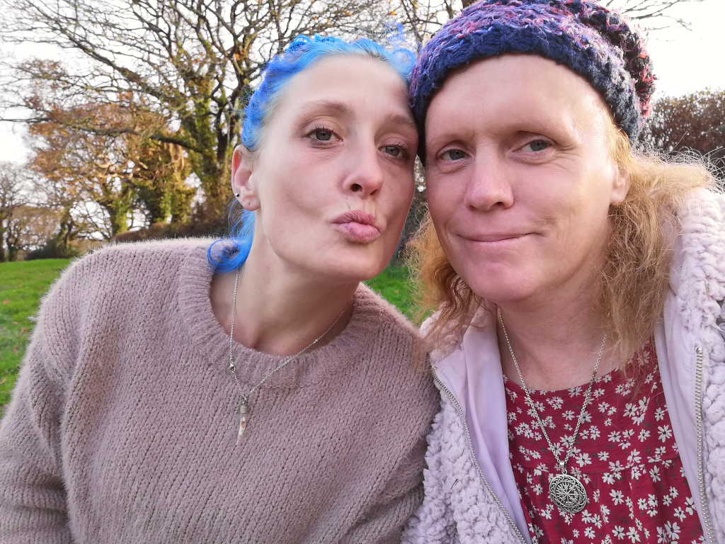 Close-up selfie of two women outdoors. The woman on the left has bright blue hair and is puckering her lips for a kiss. The woman on the right has blonde hair and is wearing a knitted hat. Both women are wearing necklaces. The background is a slightly blurred out park-like setting with trees. The overall impression is one of casual intimacy and friendship.
