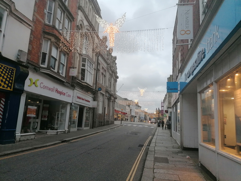 Street scene during the late afternoon or early evening. The street is lined with two-story buildings, many of which appear to be shops or businesses. Some shops display signage for charities such as Cornwall Hospice Care and the British Heart Foundation. The street is adorned with festive Christmas decorations: strings of lights and illuminated angel figures hang overhead. The overall atmosphere is quiet and peaceful, with few people visible. The sky is overcast, adding to the subdued mood. The perspective is from a point on the sidewalk, looking down the street.