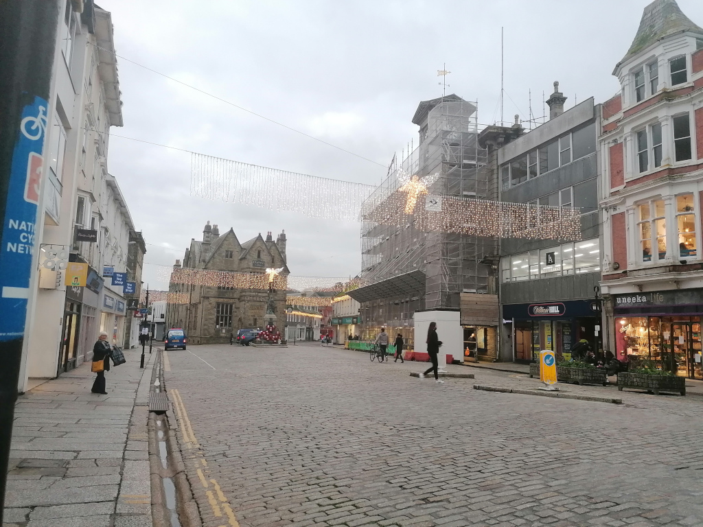 Street scene in a town or city centre, likely in the UK. The street is cobbled and relatively quiet, with a few pedestrians visible. Buildings line both sides of the street, a mix of commercial and residential properties. Christmas decorations, specifically strings of lights, hang across the street, adding a festive atmosphere despite the overcast sky. One building is under scaffolding, suggesting ongoing construction or renovation work. The overall impression is one of a peaceful, slightly subdued, yet festive town centre.