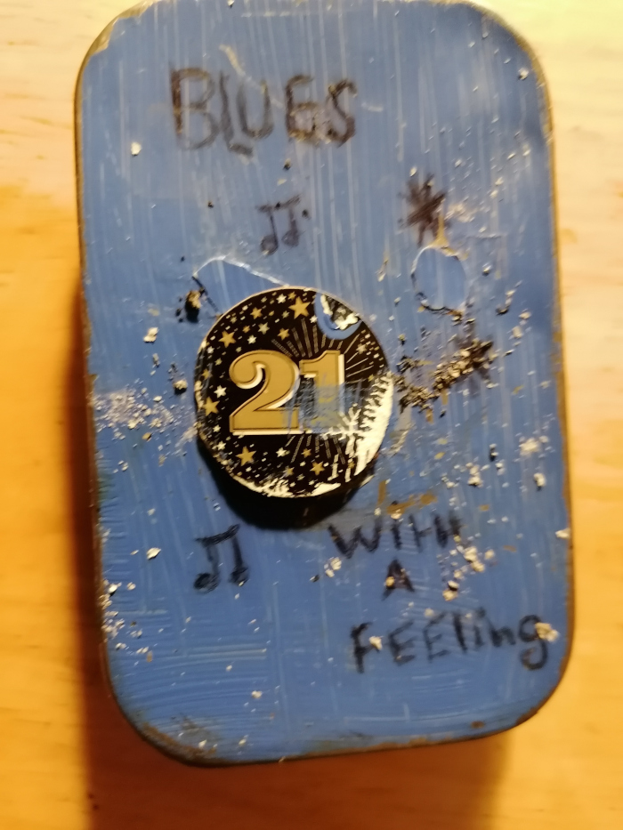 Repurposed tin, painted blue and decorated with various elements. The most prominent features are the handwritten words Blues and With a Feeling, a musical note symbol, and a circular sticker featuring the number 21 in gold against a dark background with stars. The tin shows some wear and tear, suggesting age or intentional distressing. The overall impression is one of a personalized, possibly handmade, object with a melancholic or reflective theme.