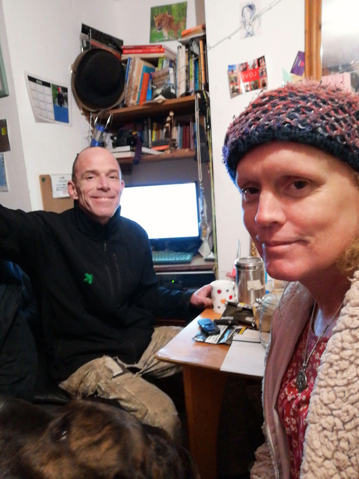 Candid shot of a man and a woman in what appears to be their home. The man is seated, wearing a dark zip-up jacket and camouflage pants. He is smiling at the camera. The woman is seated next to him and is wearing a knitted hat and a cream-coloured jacket over a red floral blouse. She looks directly at the camera with a neutral expression. A dog is partially visible in the foreground. The background is cluttered, showing bookshelves filled with books and other items, creating a lived-in and somewhat chaotic atmosphere. A computer monitor is visible behind the couple. The overall impression is one of intimacy and everyday life within a cosy but disorganised space.