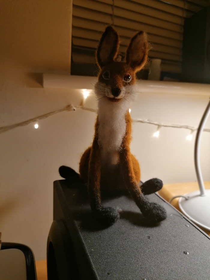 Needle-felted fox figurine sitting on top of a black speaker. The fox is predominantly orange-brown with white fur on its chest and black paws. It has a cheerful expression. The background is dimly lit, with a string of fairy lights visible behind the fox. The overall atmosphere is warm and cosy.