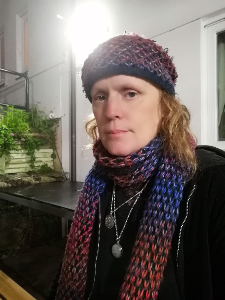 Close-up portrait of a fair-skinned woman with shoulder-length reddish-blonde hair. She, Leonie, is wearing a dark-coloured knitted beanie, a multicoloured knitted scarf (primarily shades of red, blue, and purple), and a black jacket. She also has two necklaces with circular pendants. The background is blurry but suggests an outdoor setting at night, possibly a patio or balcony, with some greenery visible. The woman's expression is neutral to slightly sombre.