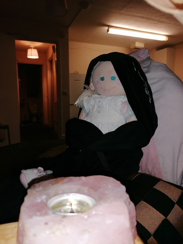 Rosie rag doll, seemingly homemade or vintage, with pale skin, large teal eyes, and rosy cheeks, dressed in a white dress. The doll is nestled in a dark-coloured hooded garment, possibly a jacket or shawl, which obscures most of its body. The setting appears dimly lit, with the doll situated near a lit pink candle resting on what looks like a rough-hewn stone or mineral. The background is blurry but suggests a sparsely furnished room. The overall ambience is subdued, mysterious, and somewhat melancholic.