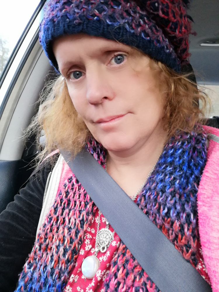 Selfie of a Leonie sitting in a car. She is wearing a blue and red knitted hat and scarf, and a red top with small white flowers. She also wears two necklaces, one with a large moonstone pendant and the other with a circular metallic pendant. Her expression is somewhat serious or pensive. The car's interior and a part of the outside are visible in the background.