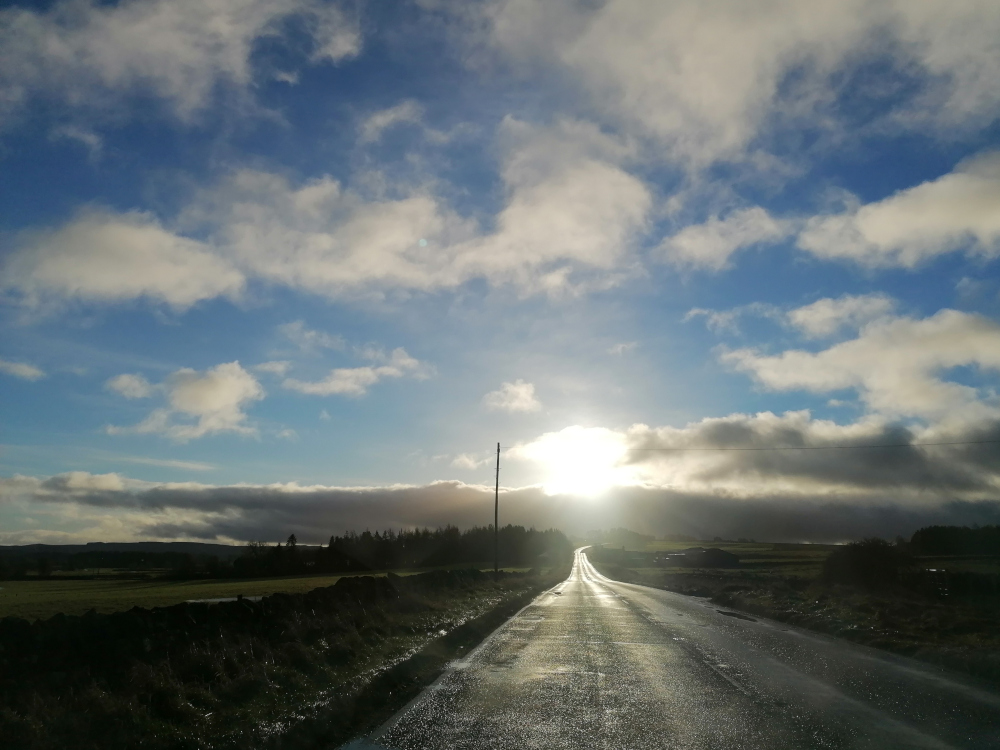 Long, straight road stretching towards a bright sun in a partly cloudy sky. The road is wet, reflecting the light. The surrounding landscape is flat and appears rural, with fields and a line of trees in the distance. The overall mood is serene and slightly melancholic, with a sense of openness and journey.
