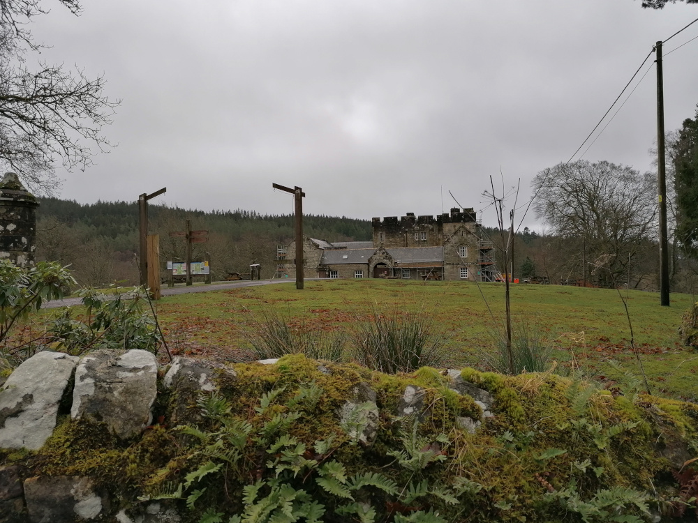 Stone castle-like building under a cloudy sky. The building appears to be undergoing some form of renovation or maintenance, as scaffolding is visible. In the foreground is a low stone wall covered in moss and vegetation, with a grassy field separating the wall and the building. There are also wooden signage posts and a utility pole visible. The overall impression is one of a somewhat secluded and possibly historic site in a rural setting.