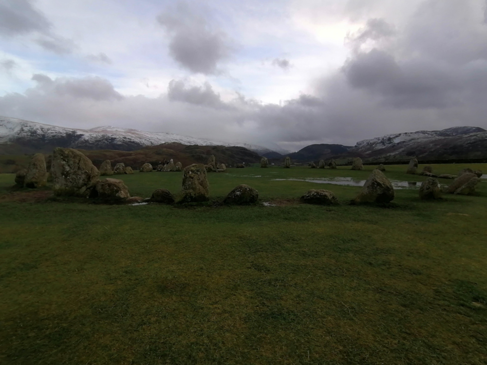 Stone circle situated in a grassy field, with snow-capped mountains in the background under a cloudy sky. The stone circle appears ancient and weathered, and some puddles are visible on the ground within the circle. The overall mood is somber and somewhat atmospheric.