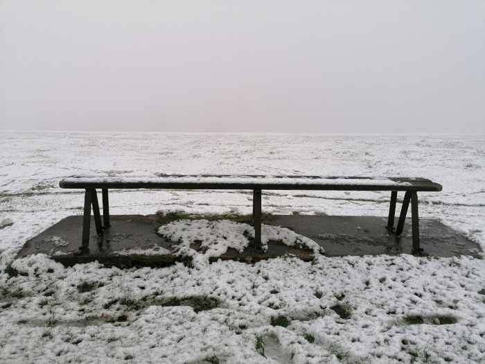 Long, dark bench covered in a light layer of snow, sitting on a concrete slab. The bench is situated in a snow-covered, open field under a foggy, overcast sky. The overall mood is bleak and desolate. There is a sense of emptiness and isolation conveyed by the vast, empty space and the solitary bench.