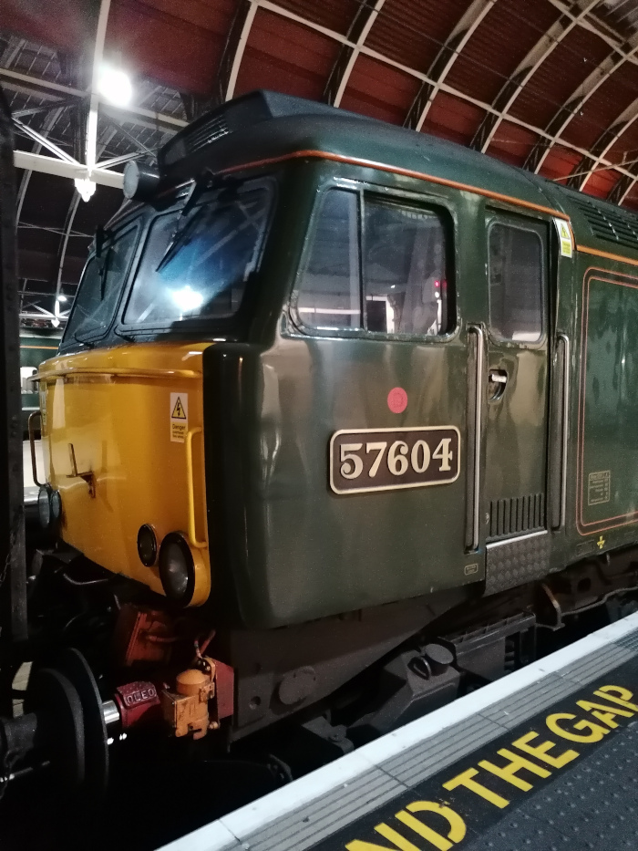 Front and side of a British Rail Class 57 locomotive, number 57604. The locomotive is predominantly dark green with a yellow front end. It's situated in what appears to be an indoor train station, under a curved metal roof. The number is clearly visible on a brass plate affixed to the side.