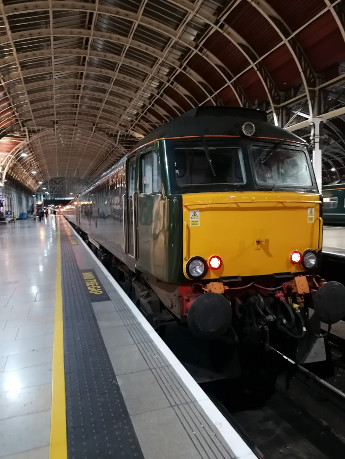 British Rail Class 57 diesel locomotive, predominantly dark green with a yellow front, standing at a train platform. The train is partially visible, suggesting it's part of a longer passenger train. The platform is located within a large, arched train station with an ornate, metallic ceiling structure. The overall mood is calm and somewhat nostalgic due to the classic design of the locomotive and the architecture of the station.