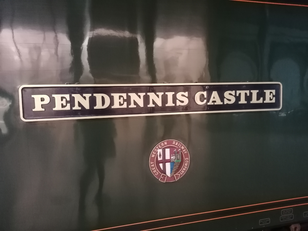 Close-up view of a dark green train car. A rectangular brass nameplate affixed to the side reads PENDENNIS CASTLE in off-white lettering. Below the nameplate is a circular emblem featuring the Great Western Railway Company logo.