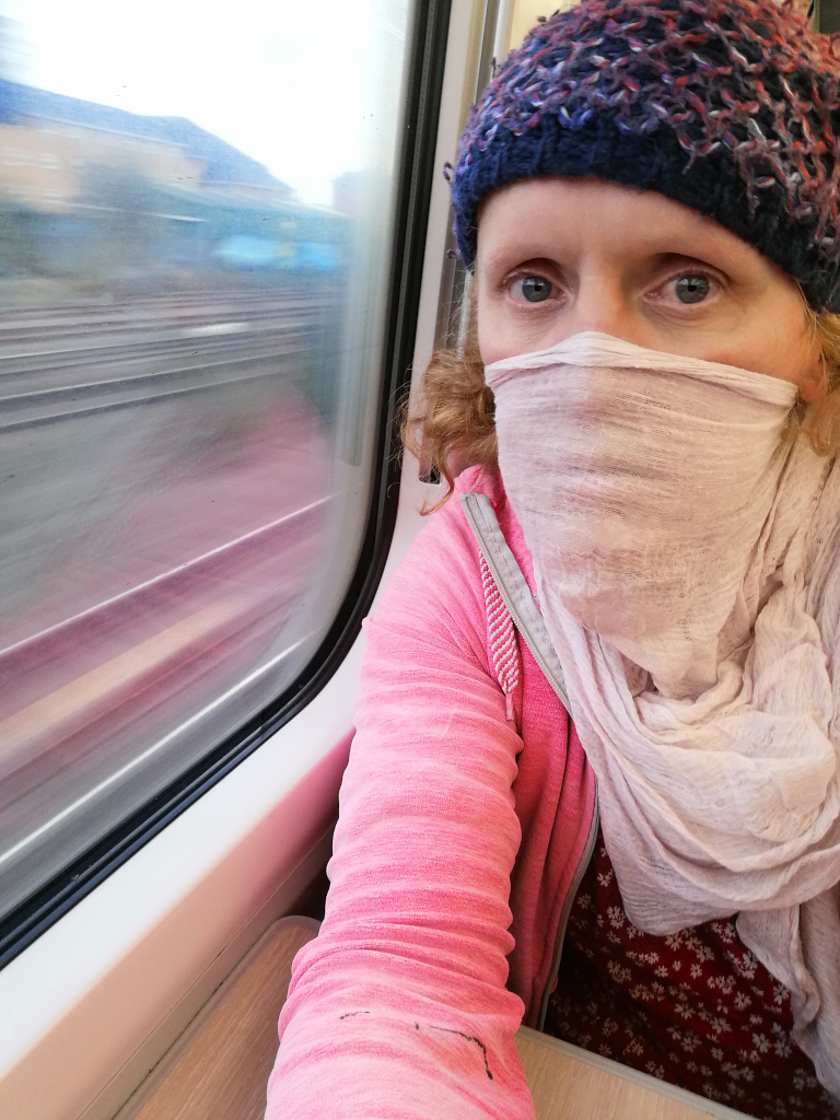 Close-up selfie of a woman on a train. She's wearing a pink sweater, a patterned dress underneath, a knitted hat, and a light-coloured scarf or shawl covering her mouth and nose. The train window, showing a blurred landscape and tracks indicating the train is moving, is visible in the background. The woman’s expression is partially obscured by the scarf, making it difficult to read her emotion fully.