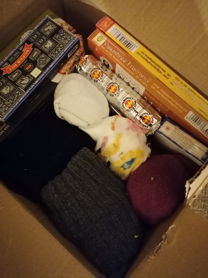 Inside of a cardboard box containing various items. Most prominently are several packages of incense sticks (Satya Nag Champa and Vijayshree Fragrance brands), and a box of what appears to be incense charcoal. There are also several pairs of socks, some appearing new and some worn or damaged, in various colours (grey, dark blue, burgundy). The overall impression is of a care package or a box of miscellaneous personal items.