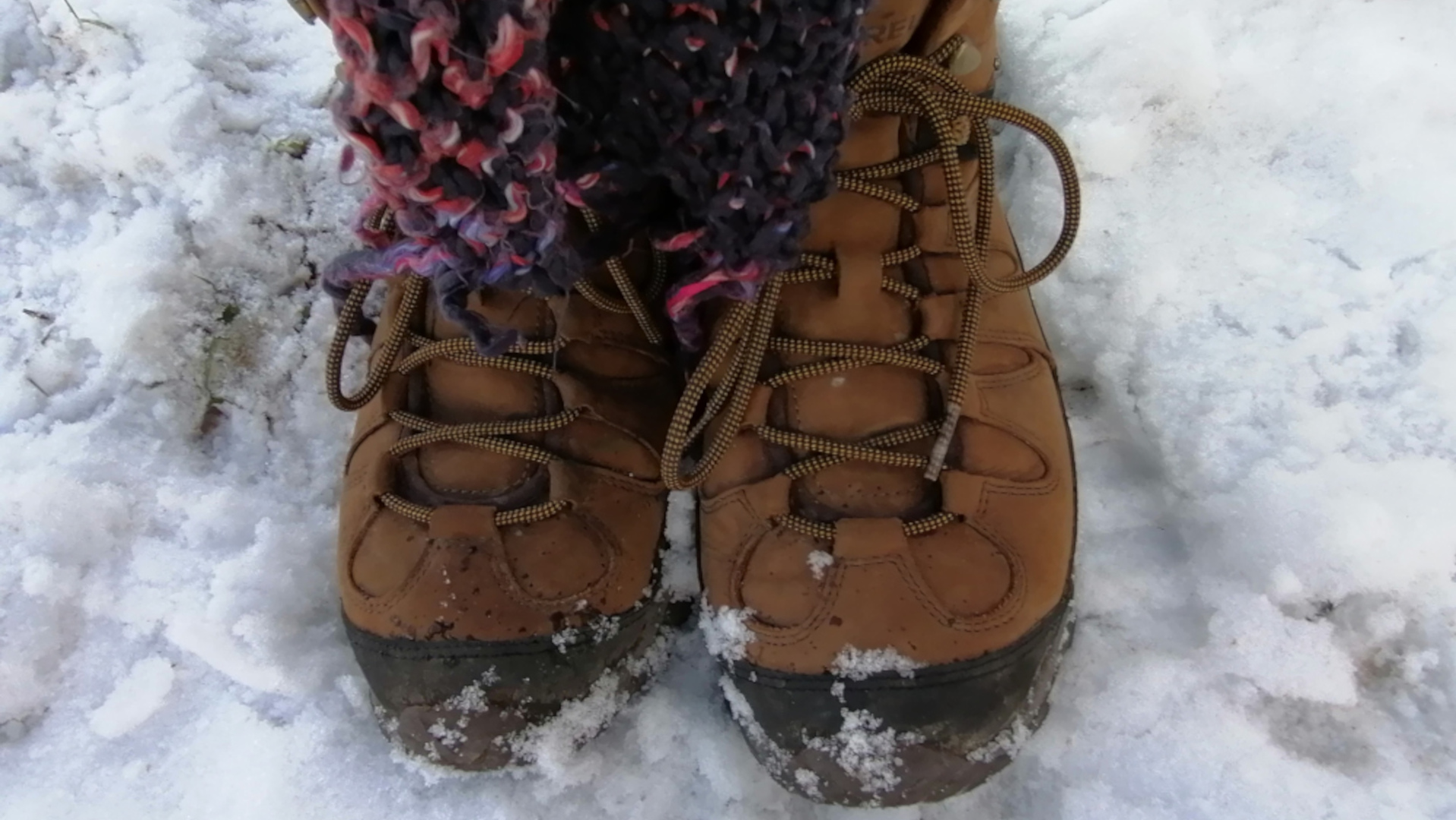 A close-up view of Leonie's feet clad in brown leather hiking boots, standing on a snowy ground. The boots are laced up and appear sturdy, suitable for winter conditions. A portion of a chunky, multicoloured knit scarf is visible wrapped around the ankles. The snow is lightly covering the ground, with some patches appearing denser than others. The overall impression is one of cold weather and outdoor activity.