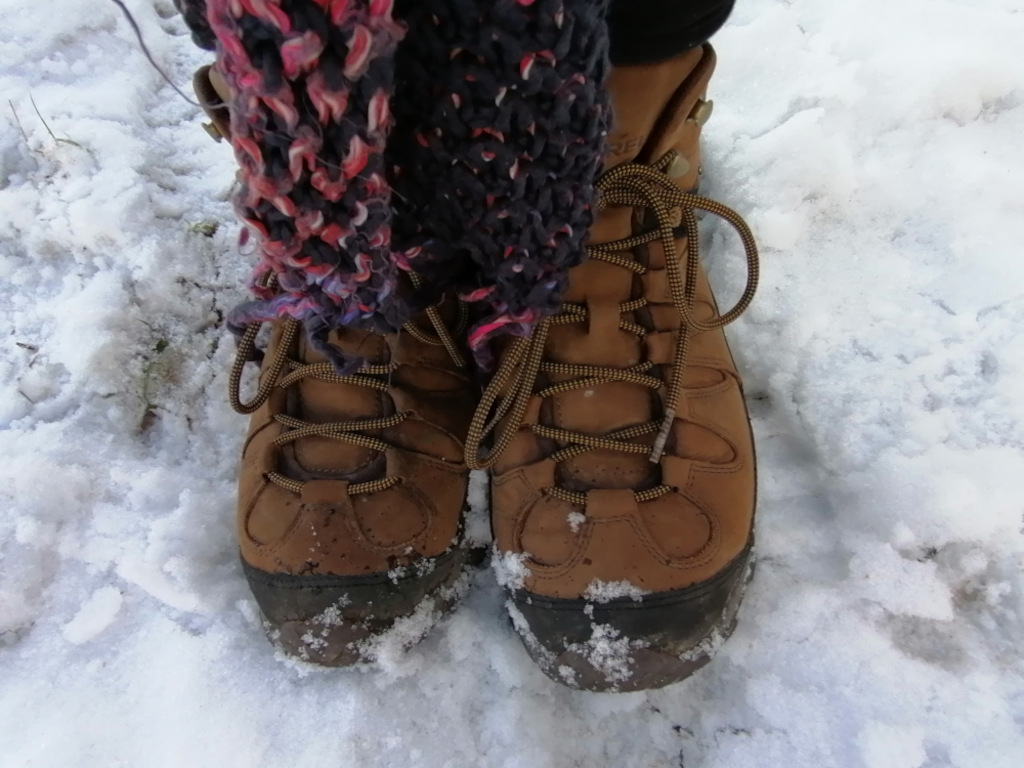 Close-up view of Leonie's feet clad in brown leather hiking boots, standing on a snowy ground. The boots are laced up and appear sturdy, suitable for winter conditions. A portion of a chunky, multicoloured knit scarf is visible wrapped around the ankles. The snow is lightly covering the ground, with some patches appearing denser than others. The overall impression is one of cold weather and outdoor activity.