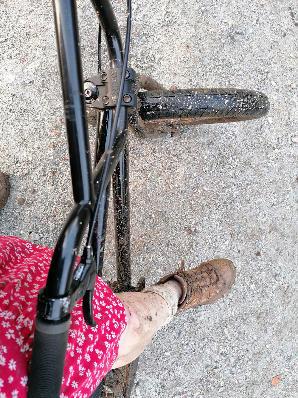 First-person perspective of a Leonie's legs and a BMX bike. Leonie is wearing a red floral dress and brown hiking boots. Their legs and the bike are covered in mud, suggesting they've been riding off-road. The focus is on the bike's handlebars and front wheel, which are also muddy. The background is a light grey gravel surface. The overall impression is one of a muddy, outdoor biking adventure.