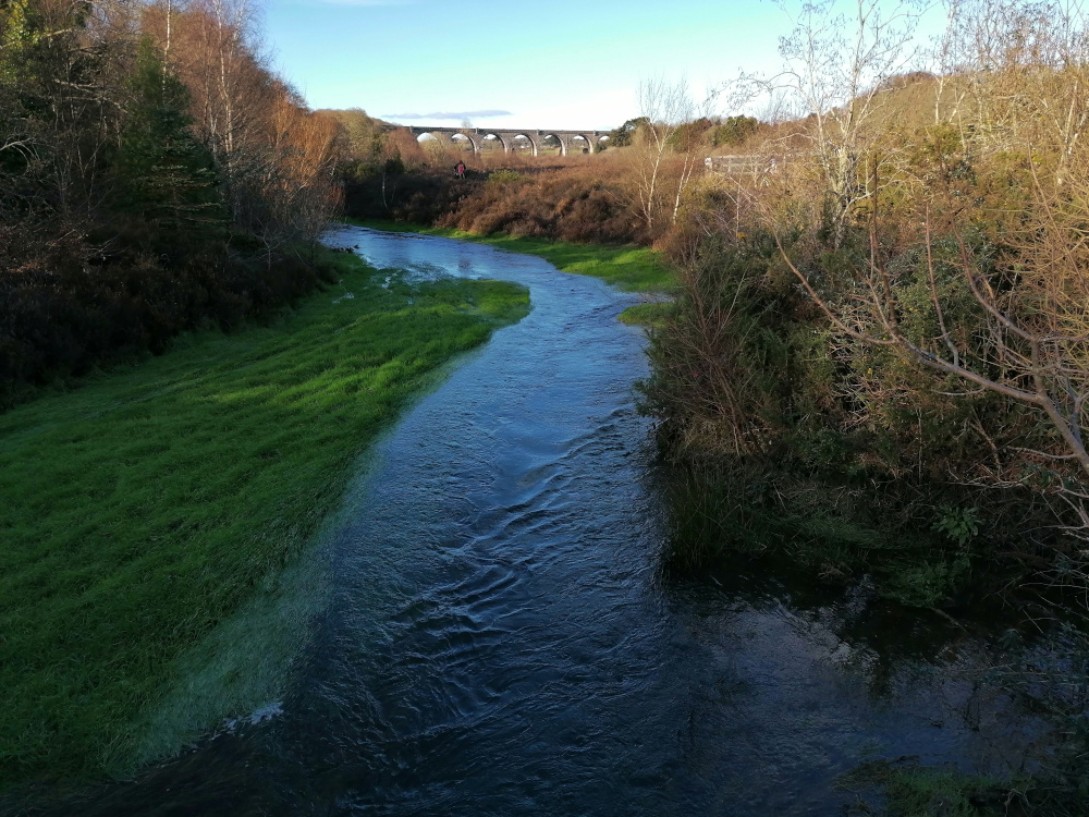 Tranquil river scene. A gently flowing stream meanders through lush green grass that lines its banks. In the distance, a stone viaduct or bridge is visible, suggesting a sense of history and perhaps industrial or transportation past. The overall mood is peaceful and serene, indicative of a natural setting, possibly in a rural area. The sky is clear and blue, suggesting daytime.