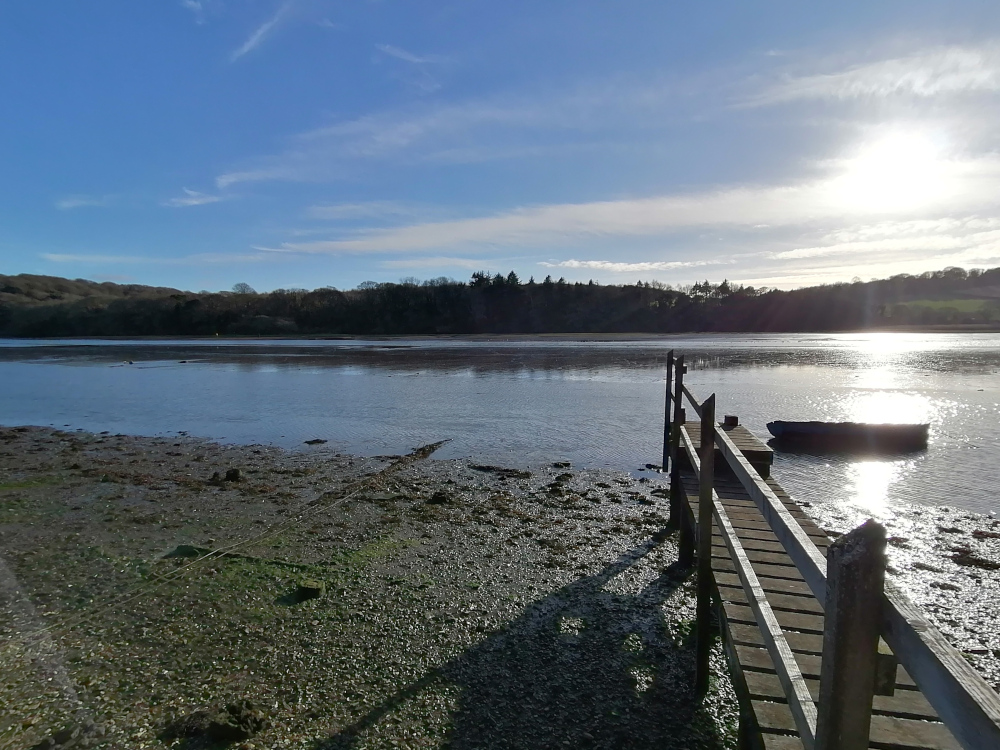 Simple wooden jetty extends from the muddy shore into the water. In the distance, the opposite bank is lined with trees and hills under a mostly clear, sunny sky. A small, dark-coloured boat is moored near the jetty. 