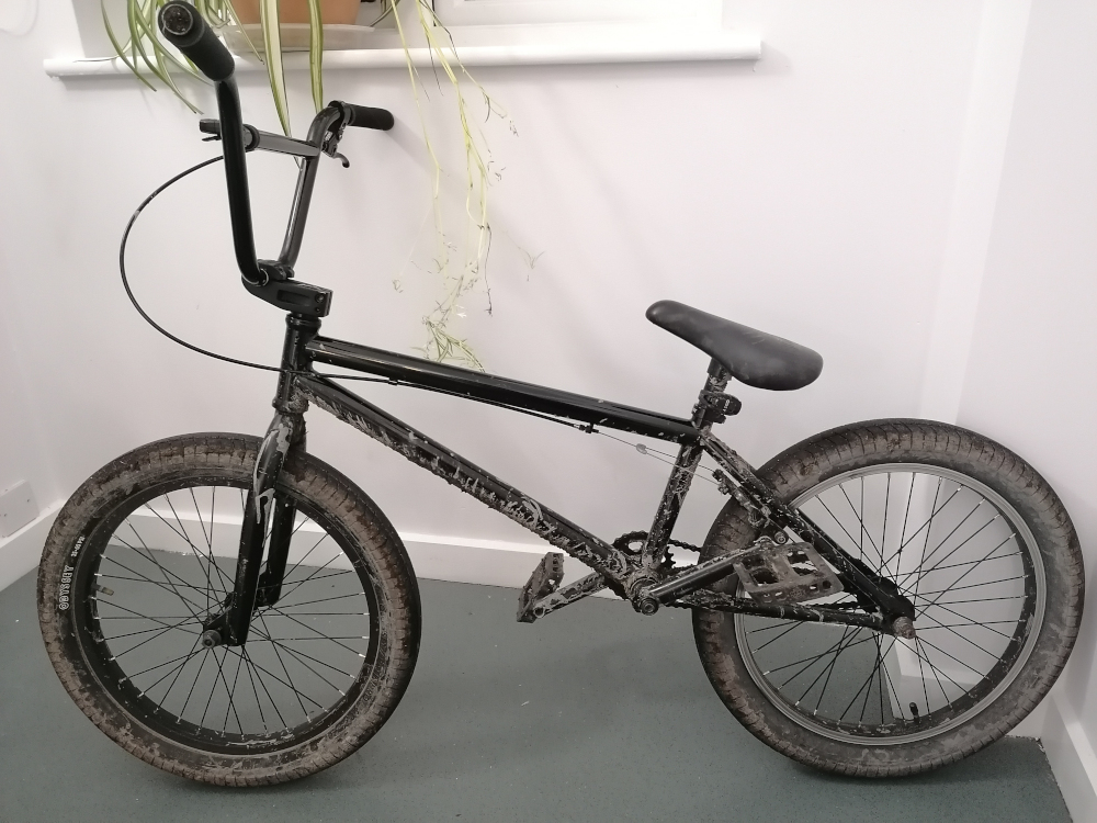 Black BMX bicycle. The bike is slightly dirty, indicating some prior use. It is positioned against a white wall. A houseplant is partially visible in the background.
