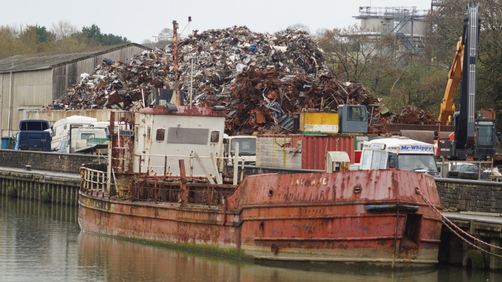 Rusty, old barge docked next to a large pile of scrap metal. Behind the barge and scrap pile are several trucks, containers, and industrial-sized machinery, suggesting a scrap yard or recycling facility located on a waterway. The overall impression is one of industrial decay and the accumulation of discarded materials.
