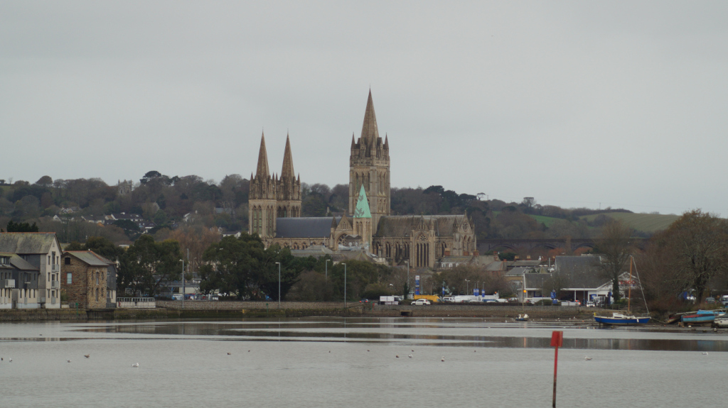 Truro Cathedral in Cornwall, England, as seen from across a body of water.  The cathedral is prominent in the mid-ground, its tall spires and stonework visible against a muted, overcast sky.  In the foreground, there's calm water, a few birds, and a small boat. The immediate foreground shows the calm water reflecting the sky. Buildings and trees are visible in the background, along the shore. The overall mood is tranquil and slightly sombre due to the grey sky.