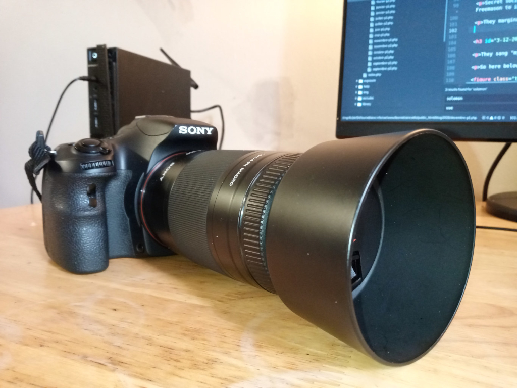 Sony Alpha DSLR camera with a large lens attached and a lens hood. The camera is positioned on a light-brown wooden surface. A small black device (possibly a wireless transmitter) is attached to the camera's hot shoe. In the background, a computer monitor displays code. The overall impression is that of a photographer's workspace or setup, possibly involved in some form of digital photography and/or post-processing workflow judging by the visible code. There is no discernible narrative or deeper moral message beyond a straightforward depiction of photographic equipment.