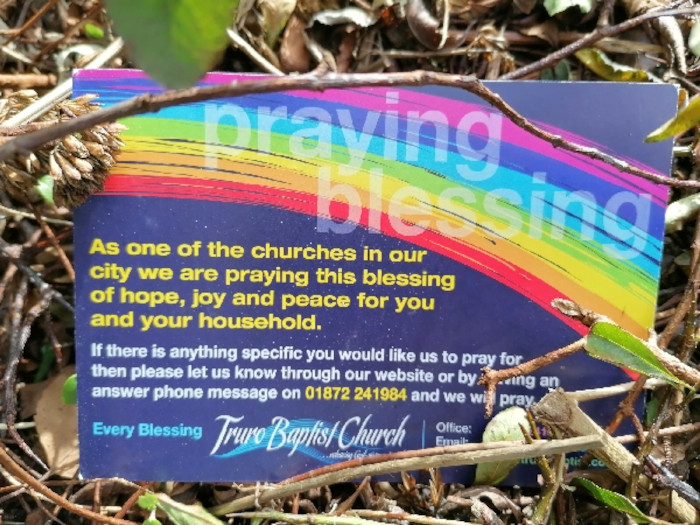 Card lying on the ground amidst fallen leaves and twigs. The card is a dark blue-purple, features a rainbow arc, and contains a message from Truro Baptist Church. The message states that the church is praying a blessing of hope, joy, and peace for the recipient and their household. It also invites people to request specific prayers through their website or by leaving a phone message. The overall feeling is one of offering comfort, support, and spiritual outreach. The card's placement on the ground suggests a random act of kindness or a broader community outreach initiative.
